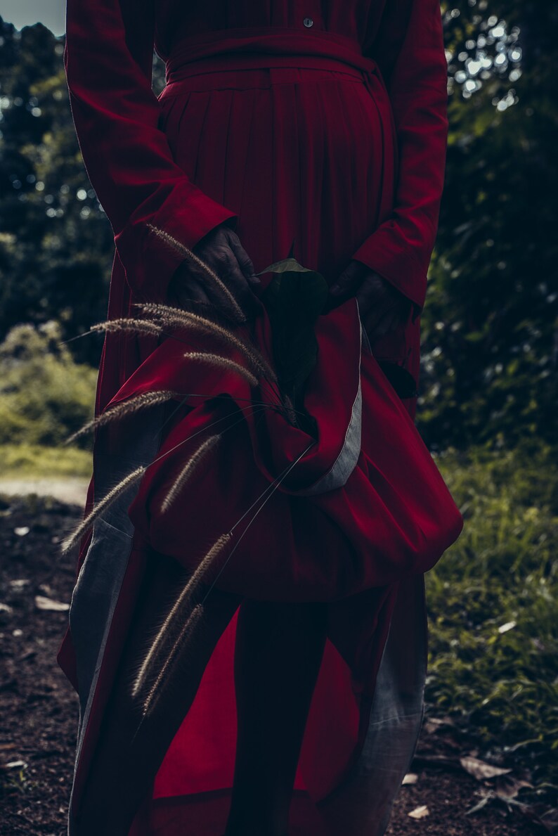 May include: A person wearing a long, red dress with a white belt. The dress is flowing and has a loose fit. The person is standing in a forest setting with tall grass and trees in the background.