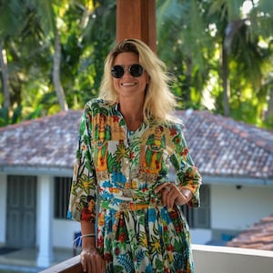 May include: A woman wearing a white, green, and blue floral print dress with a long sleeve and a belt. She is smiling and wearing round sunglasses.