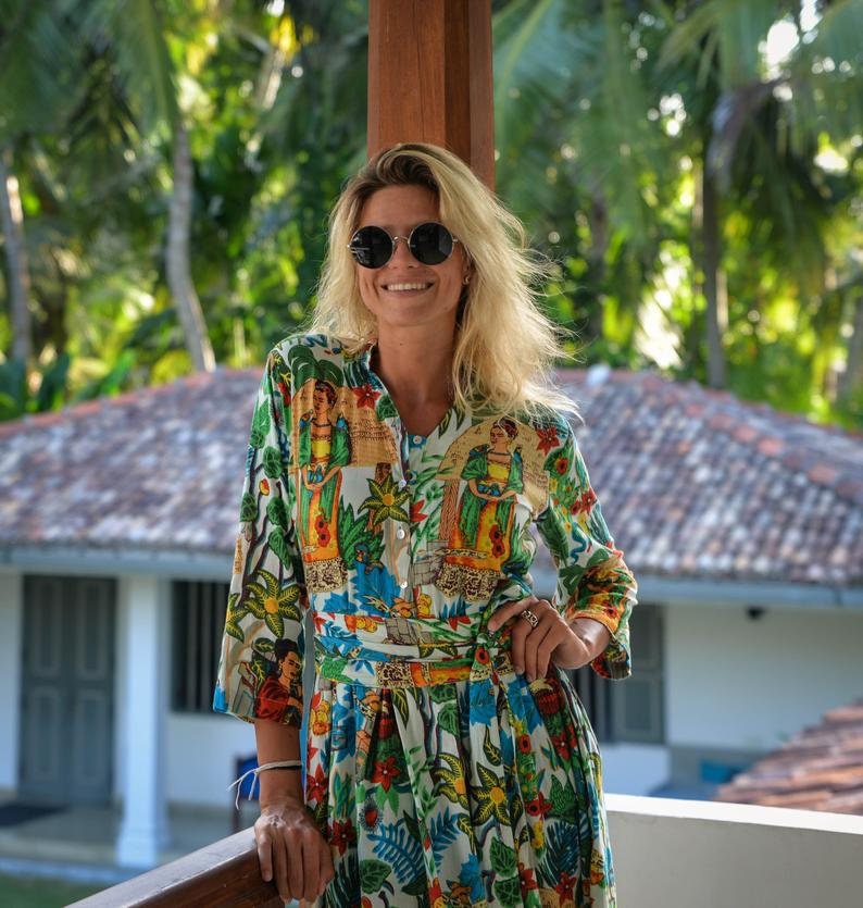 May include: A woman wearing a white, green, and blue floral print dress with a long sleeve and a belt. She is smiling and wearing round sunglasses.