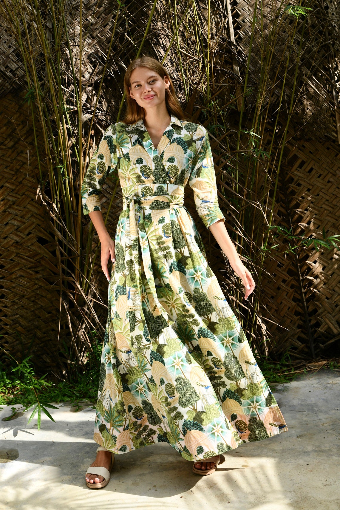 May include: A long, white dress with a green and blue floral print. The dress has a wrap style and a tie at the waist. The model is wearing the dress with white sandals.
