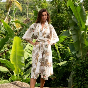 May include: A white kimono robe with a floral print and gold accents. The robe has a tie belt and is worn by a person standing in a tropical garden.