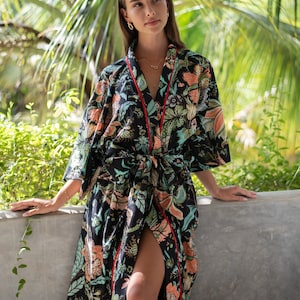 Pode incluir: Uma bata de kimono preta com estampa floral, detalhes vermelhos e cinto de amarrar. A bata é usada por uma pessoa em pé em frente a um fundo verde frondoso.