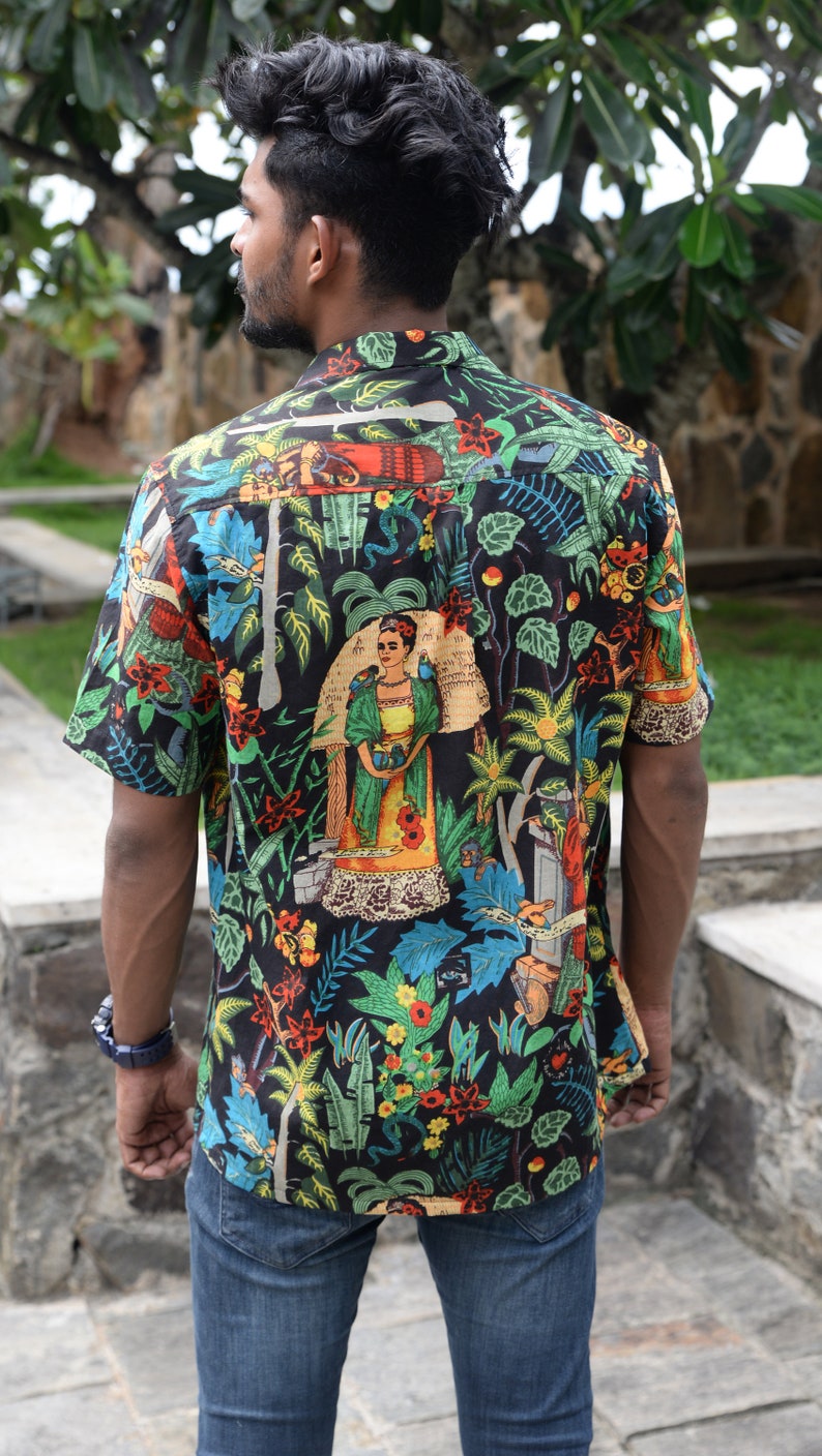 May include: Short-sleeved button-down shirt with a vibrant, colorful print. The black shirt features a botanical design with images of a woman, plants, and animals. The shirt is paired with blue jeans.