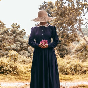 May include: A woman wearing a wide-brimmed straw hat and a long, dark blue dress holds a bouquet of pink flowers. She is standing on a path in a forest setting.