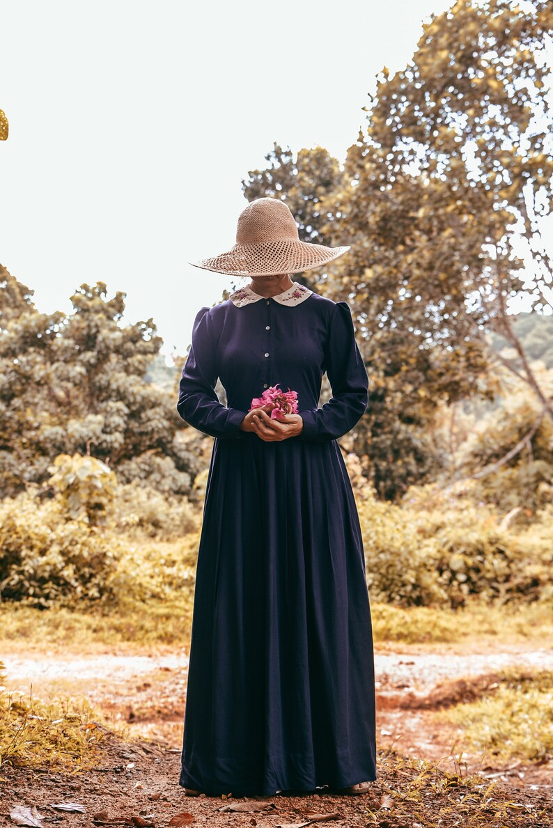 May include: A woman wearing a wide-brimmed straw hat and a long, dark blue dress holds a bouquet of pink flowers. She is standing on a path in a forest setting.