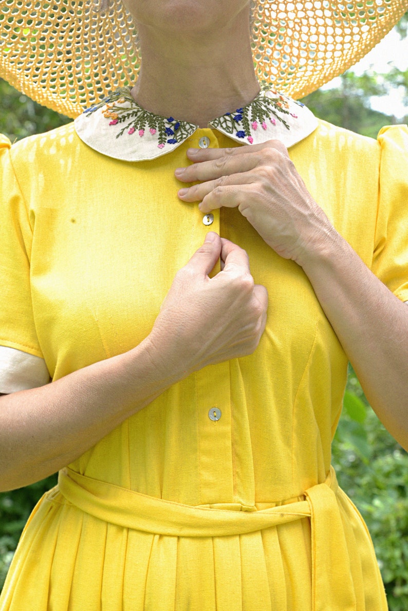 May include: A woman wearing a yellow dress with a white collar embroidered with flowers. She is fastening a button on the front of the dress.