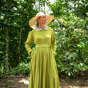 May include: A woman wearing a long, lime green dress with a white floral collar and a straw hat. She is standing in a forest setting.