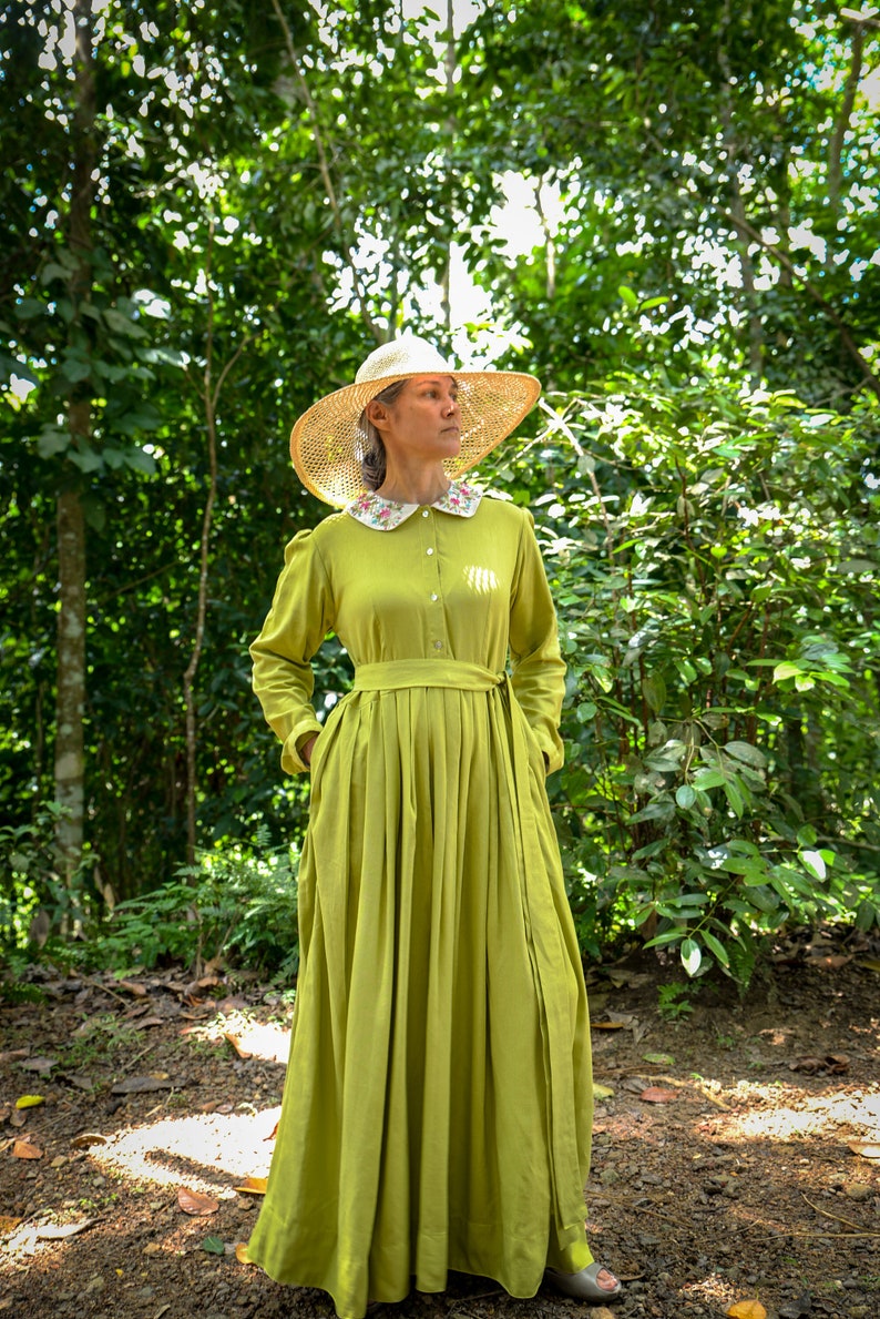 May include: A woman wearing a long, lime green dress with a white floral collar and a straw hat. She is standing in a forest setting.