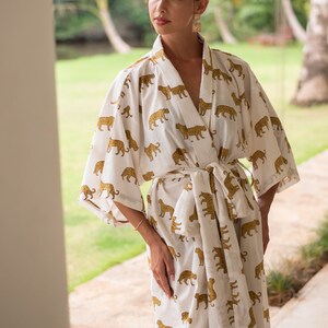 May include: A white robe with a gold leopard print. The robe has long sleeves and a tie belt. The model is wearing gold earrings.