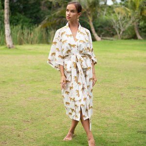 May include: A white robe with a gold tiger print. The robe has long sleeves and a tie belt. The model is standing on a grassy lawn.