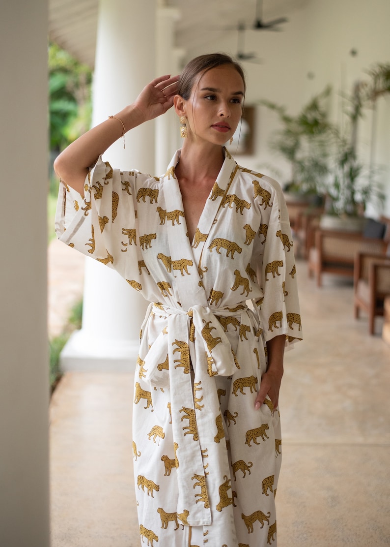 May include: A white robe with a gold leopard print. The robe has long sleeves and a tie belt. The robe is being worn by a person with long brown hair and is standing in a white room.