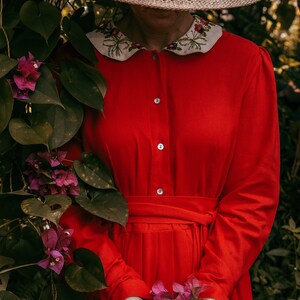 May include: A woman wearing a red dress with a white embroidered collar and a wide-brimmed straw hat holds a bouquet of pink flowers.