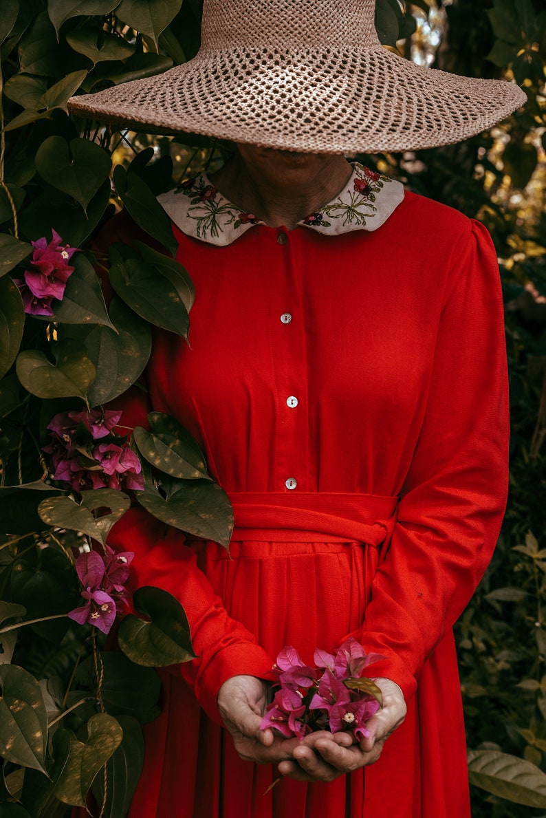 May include: A woman wearing a red dress with a white embroidered collar and a wide-brimmed straw hat holds a bouquet of pink flowers.