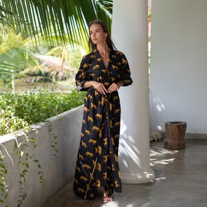May include: A woman wearing a long black robe with a gold animal print. The robe has long sleeves and a tie belt. The woman is standing in a room with white walls and a wooden floor.
