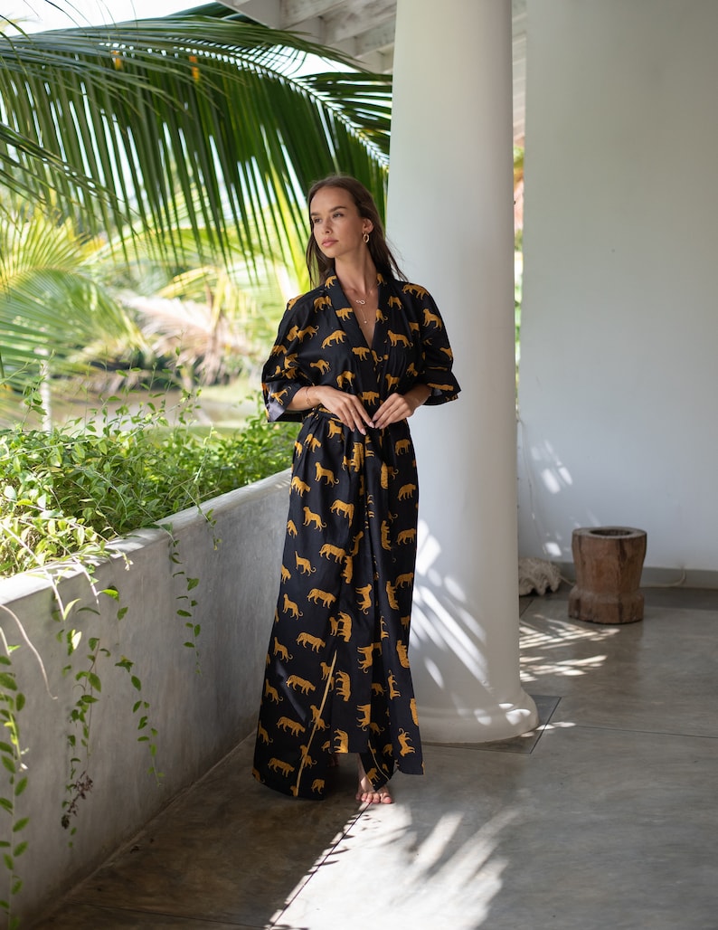 May include: A woman wearing a long black robe with a gold animal print. The robe has long sleeves and a tie belt. The woman is standing in a room with white walls and a wooden floor.