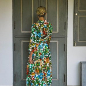 May include: A woman wearing a long, white dress with a colorful floral and figural print. The dress has a cinched waist and a flowing skirt. She is standing in front of a closed wooden door.