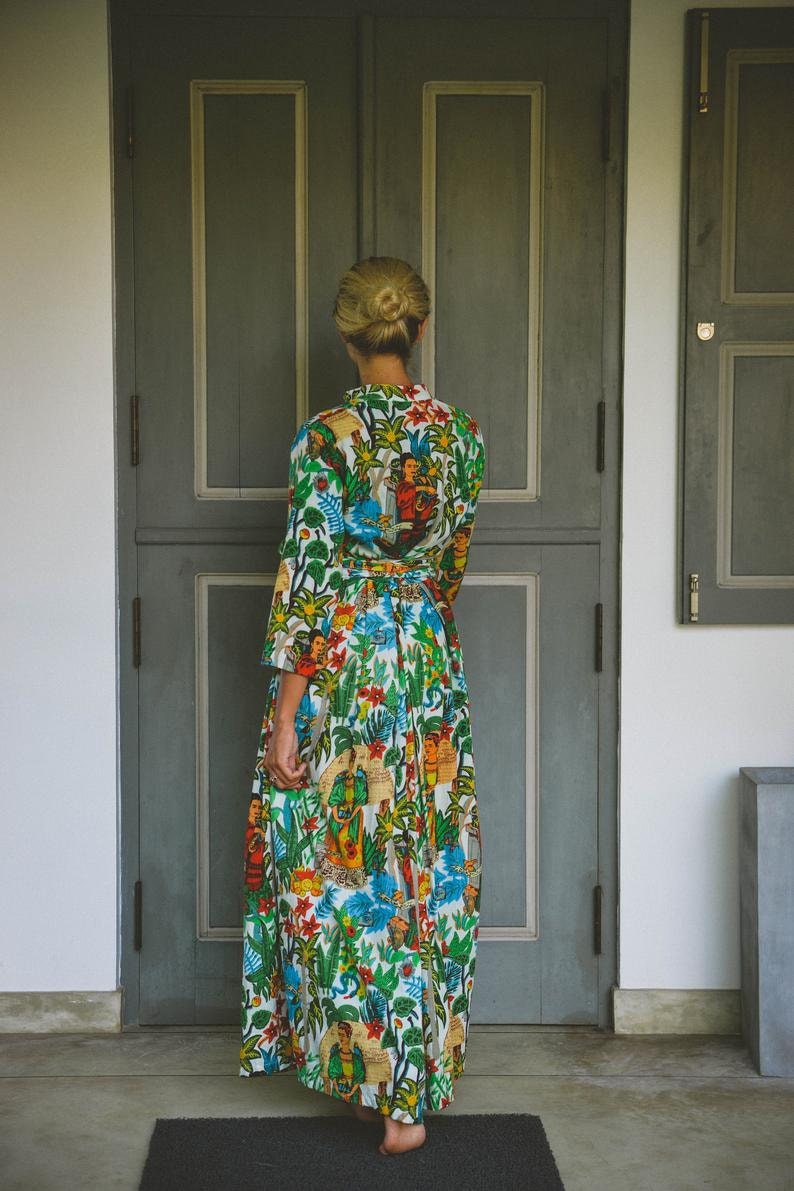 May include: A woman wearing a long, white dress with a colorful floral and figural print. The dress has a cinched waist and a flowing skirt. She is standing in front of a closed wooden door.