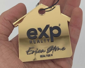 Op de afbeelding: Een gouden sleutelhanger in de vorm van een huis met de tekst "exp realty" en "Erica Stone Realtor®" in zwarte letters.