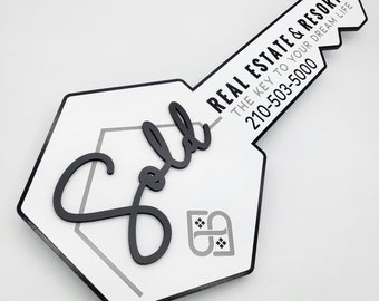 May include: A white and black key-shaped sign with the text "REAL ESTATE & RESORTS THE KEY TO YOUR DREAM LIFE 210-503-5000" and the word "Sold" in a black cursive font.