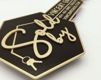 Könnte beinhalten: Ein schwarzes und goldenes schildförmiges Schlüssel-Schild mit dem Text "Verkauft von" in goldenen Buchstaben und den Kontaktinformationen für Vincent Giles, Immobilienmakler. Das Schild ist als Immobilien-„Verkauft“-Schild konzipiert.