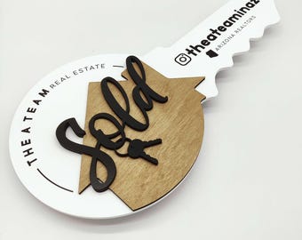 May include: A white round sign with black text that says "The A Team Real Estate" and a black key with a house shape and the word "Sold" in black script. The key is attached to a white key tag with black text that says "@theateaminaz ARIZONA REALTORS".