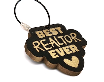 Op de afbeelding: Een zwarte en gouden sleutelhanger met de tekst "BEST REALTOR EVER" en een hartsymbool. De sleutelhanger heeft een zwarte draadlus en een zilveren sluiting. De sleutelhanger heeft de vorm van een klavertjevier.