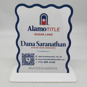May include: A white and blue acrylic business card holder with the Alamo Title logo and contact information for Dana Saranathan. The holder has a wavy blue border and a white base. The text includes a QR code and the hashtag #DAZZLEwithDANA.