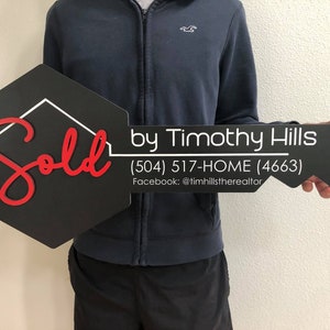 May include: Black and white key-shaped sign with the word "Sold" in red cursive lettering. The sign also includes the text "by Timothy Hills (504) 517-HOME (4663) Facebook: @timhillstherealtor".