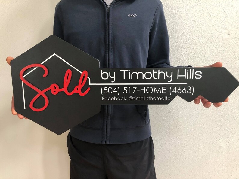 May include: Black and white key-shaped sign with the word "Sold" in red cursive lettering. The sign also includes the text "by Timothy Hills (504) 517-HOME (4663) Facebook: @timhillstherealtor".