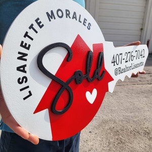 May include: A white and red wooden key-shaped sign with the word "Sold" in black script. The sign also includes the text "Lisa nette Morales" and a phone number, "407-276-7042", and the text "@RealtorLisa nette".