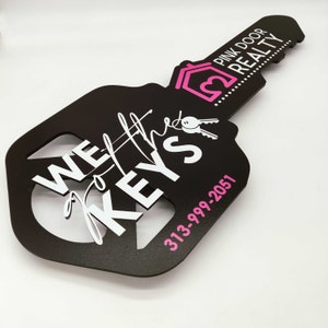 Pode incluir: Um sinal preto em forma de chave com o texto "We've got the keys" em letras brancas. O sinal também apresenta um ícone de casa rosa com o texto "Pink Door Realty" e um número de telefone: 313-999-2051.