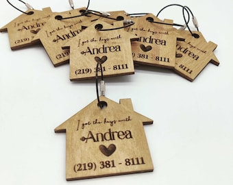 Op de afbeelding: Houten sleutelhangers in de vorm van een huis met de tekst "Ik heb de sleutels met Andrea" en een telefoonnummer. De sleutelhangers hebben een hartvormig ontwerp en zijn bevestigd aan een zwart koord.