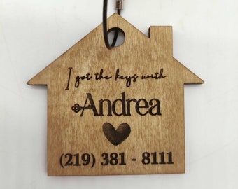 Op de afbeelding: Houten sleutelhanger in de vorm van een huis met de tekst "Ik heb de sleutels met Andrea" en een hartje, samen met een telefoonnummer (219) 381-8111.