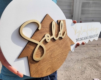 Puede incluir: Un cartel de madera blanco y marrón en forma de llave con la palabra "Vendido" en letras doradas. El cartel también incluye el texto "Kylie Frederickson 707.567.6361 | REALTOR*" en letras doradas.