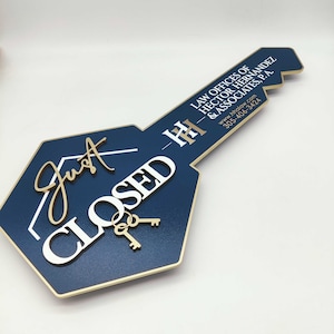 Può includere: Un cartello a forma di chiave blu e dorato con il testo "Just Closed" in lettere bianche e dorate. Il cartello include anche il testo "Law Offices of Hector Hernandez & Associates, P.A." in lettere nere, insieme a un indirizzo di sito web e un numero di telefono.
