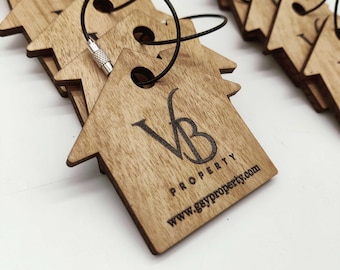 Op de afbeelding: Een stapel houten sleutelhangers in de vorm van een huis met een zwarte touwlus. De sleutelhanger heeft de tekst "VB PROPERTY" en de website "www.gsyproperty.com".