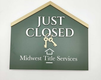 May include: A green sign with a gold border shaped like a house. The sign reads "JUST CLOSED" with two gold keys hanging below the text. The sign also reads "Midwest Title Services" at the bottom.
