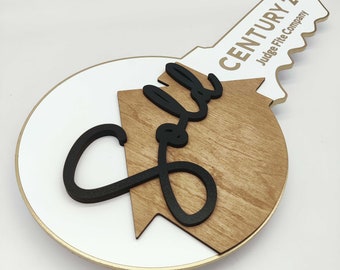 May include: A wooden key-shaped sign with a white circle and a brown house shape with the word "Sold" in black script. The key has the text "CENTURY 21® Judge Fite Company" in gold lettering.