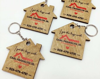 Op de afbeelding: Vier houten sleutelhangers in de vorm van een huis met metalen sleutelringen. Elke sleutelhanger heeft de tekst "I got the keys with CARL FRANKLYN JR. MY FAVORITE REALTOR - 929-479-4791-" in zwart en rood. De sleutelhangers zijn gemaakt van lichtgekleurd hout.