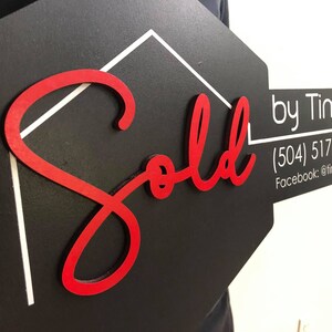 May include: A black and white house-shaped sign with a red "Sold" inscription. The sign is held by a person's hand. The sign also includes the text "by Timothy Hills" and contact information: (504) 517-HOME (466) and Facebook: @timhillstherealtor.