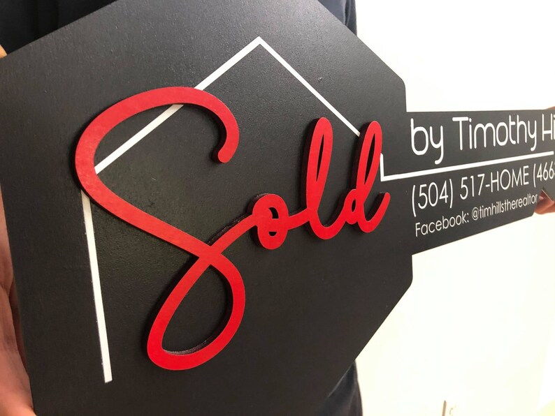 May include: A black and white house-shaped sign with a red "Sold" inscription. The sign is held by a person's hand. The sign also includes the text "by Timothy Hills" and contact information: (504) 517-HOME (466) and Facebook: @timhillstherealtor.