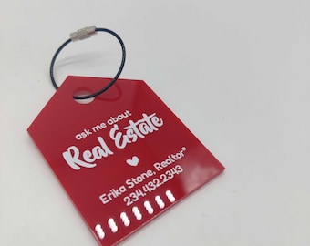 Op de afbeelding: Rode acryl sleutelhanger met een zwarte metalen ring. De sleutelhanger heeft witte tekst die "ask me about Real Estate" zegt met een hartje en "Erika Stone, Realtor" met een telefoonnummer.