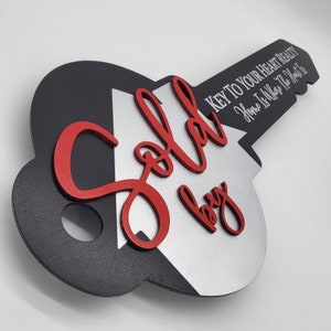 May include: A decorative wooden key-shaped sign with the words "Sold by" in red script. The key is black and silver with the text "KEY TO YOUR HEART REALTY Home Is Where The Heart Is" in white.
