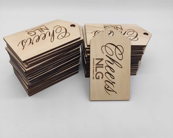 Op de afbeelding: Stapels houten cadeaulabels met een lichtbruine afwerking. De rechthoekige labels hebben een puntige bovenkant en een gat. Het woord "Cheers" staat in sierlijke letters, met "NLG Divorce & Family Law" eronder.