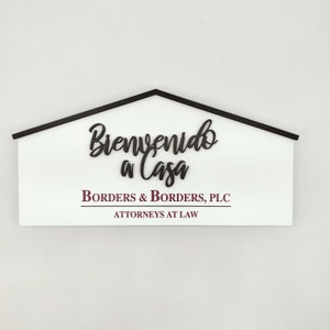 May include: A white wooden sign in the shape of a house with a black roof. The sign reads "Bienvenido a Casa" in cursive, with "Borders & Borders, PLC" and "ATTORNEYS AT LAW" below.