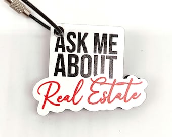 Op de afbeelding: Witte houten sleutelhanger met zwarte tekst die "Ask Me About Real Estate" in rood cursief schrift zegt. De sleutelhanger heeft een zwart koord met een zilveren metalen lus.