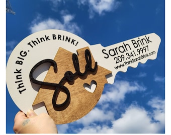 May include: A wooden real estate sign in the shape of a key, with the word "Sold" in black script. The sign also includes the text "Think BIG. Think Brink!" and contact information for Sarah Brink.