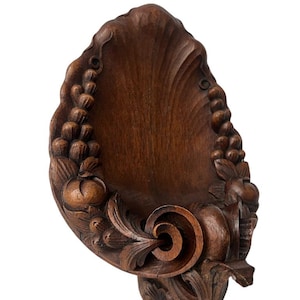 May include: A dark brown, carved wooden wall sconce with a shell-like design. The sconce features detailed carvings of grapes, leaves, and a central scroll. The overall shape is asymmetrical, with a flat back for wall mounting.