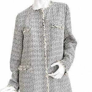 Puede incluir: Un abrigo de tweed blanco y negro con mangas largas y un dobladillo hasta la rodilla. El abrigo presenta un cuello redondo, bolsillos decorativos y ribetes blancos a lo largo de los bordes y las costuras. El abrigo se exhibe en un maniquí.