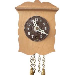 May include: A small, wooden cuckoo clock with a light brown finish. The clock face is dark brown with white numerals and hands. Decorative chains with pinecone-shaped weights hang below. The clock is a classic decorative item.